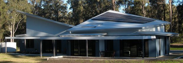 Low, single-storey modern residence set within a wooded landscape. The building has a strongly horizontal composition, broad roof planes with deep overhangs, extensive dark glazing, and corrugated or ribbed metal wall sections. A large raised central roof element and crisp geometric forms create a contemporary pavilion-like facade.