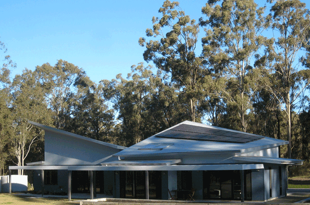 Exterior view of a low, contemporary pavilion-like house set among mature eucalyptus trees. The building has strongly articulated asymmetrical roof planes, broad shallow eaves, extensive dark glazing, slender metal posts, and corrugated metal wall cladding. The predominantly grey and charcoal facade contrasts with the surrounding bushland.