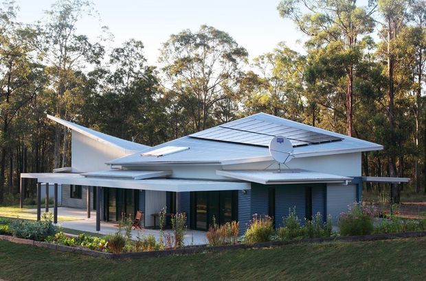 A contemporary single-storey house set within a wooded landscape. The building has a low, angular form with broad flat and skillion roof planes, pale metal roofing, dark horizontal wall cladding, extensive glazing, and covered outdoor areas supported by slender posts. Landscaped garden beds and a lawn surround the residence.