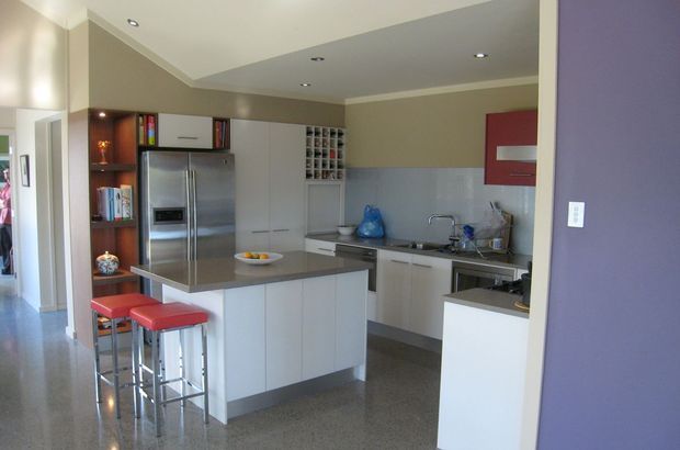 A contemporary open-plan kitchen viewed from an adjacent hallway or living area. The room features a central island with a grey stone-look benchtop and white cabinetry, two red upholstered bar stools, stainless-steel appliances, integrated pantry storage, a grey tiled splashback, recessed ceiling lights, and dark polished flooring. A purple accent wall frames the foreground.