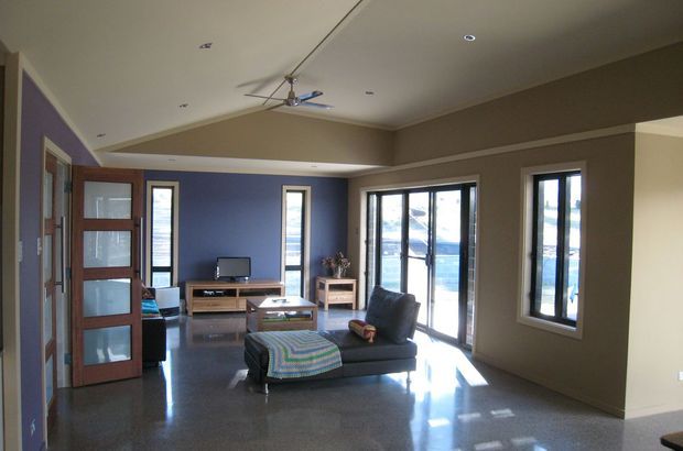 A contemporary open-plan living room with polished dark flooring, a charcoal sectional sofa and ottoman, low timber media cabinetry, and extensive glazed doors and windows. The room has taupe and deep blue walls, recessed ceiling lights, a ceiling fan, and strong natural light entering from the glazed exterior openings.