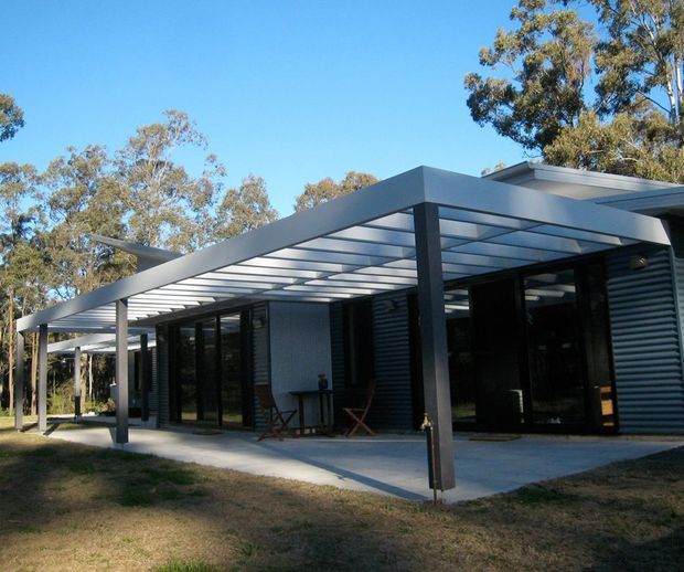 Single-storey contemporary house viewed from outside, featuring dark corrugated metal wall cladding, extensive black-framed glazing, and a large flat-roofed covered terrace or carport-like pergola with translucent roof panels and slender steel posts. The home is set among mature trees and dry landscaped ground under clear daylight.