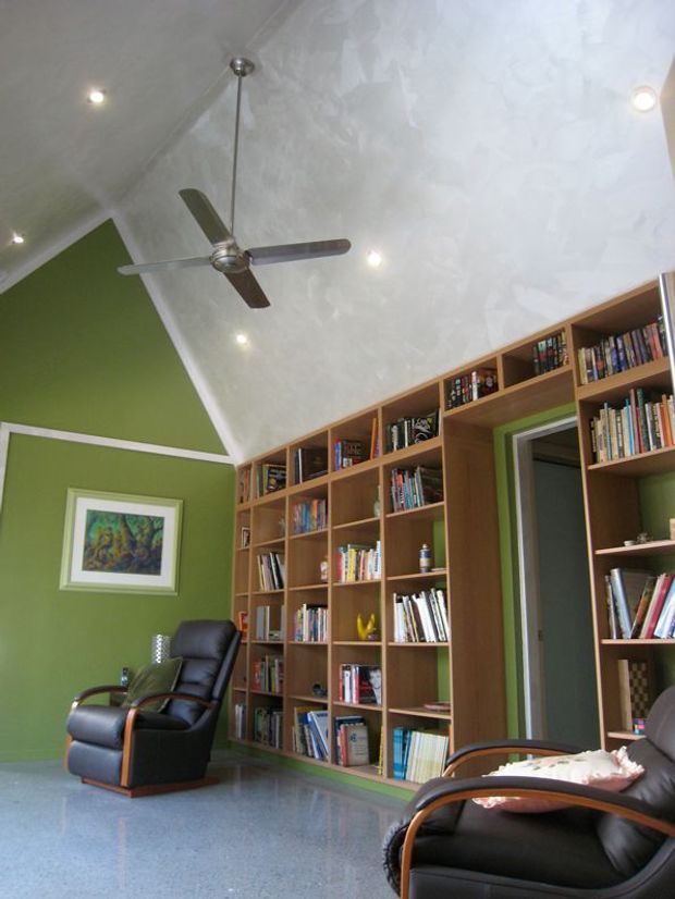 A tall, library-like reading room with floor-to-ceiling custom timber bookcases, green painted walls, a polished light grey floor, leather lounge chairs, and a large ceiling fan. The room has a sloping, textured white ceiling with recessed spotlights and an integrated doorway within the shelving. The overall impression is a calm, functional home library or living space with strong built-in joinery.