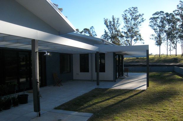 Exterior view of a low-profile modern house with overlapping flat roof planes, a broad covered patio or carport-like canopy, dark-framed glazing, and grey horizontal cladding. The house opens onto a paved terrace and lawn, with a gently sloping, tree-lined site beyond.