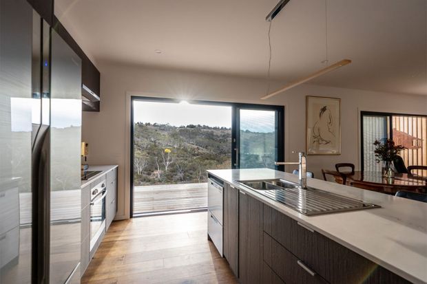 Contemporary kitchen with a large central island, integrated sink and drainer, dark textured cabinetry, pale stone benchtops, stainless-steel appliances, and warm timber-look flooring. Full-height glazing opens to a timber deck and a leafy hillside outlook. A dining area is visible beyond the kitchen.
