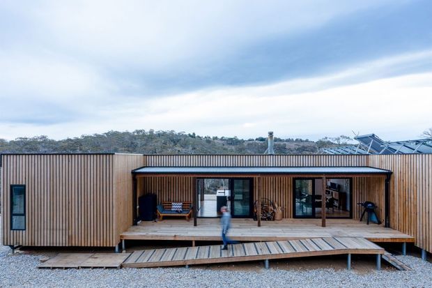 A long, single-storey contemporary timber-clad building with a deep covered veranda and broad timber deck. Full-height glazed doors open onto the deck, while a rooftop solar array and cylindrical flue are visible above the roofline. The building sits in a sparse, bushland setting beneath an overcast sky.