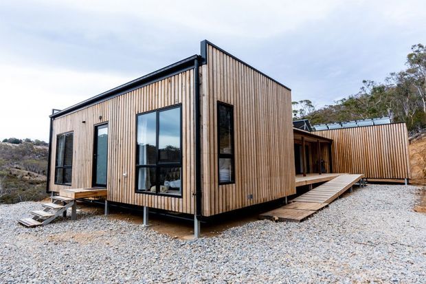 Exterior view of a compact contemporary modular or cabin-style building clad in vertical timber boards. The structure is raised on slender supports above a gravel surface, with black-framed glazing, a small timber entry deck and ramp, and a connected rear volume. It sits in a rugged, sparsely landscaped bush setting.