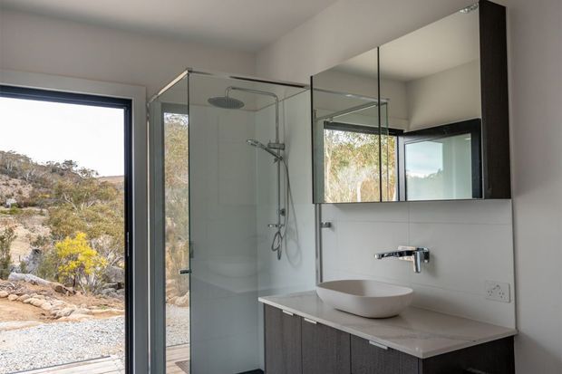 Contemporary bathroom with a frameless glass walk-in shower, wall-mounted chrome fixtures, a vessel basin on a pale stone vanity, and dark wood-look cabinetry. A large mirrored cabinet reflects the bright, minimalist interior and surrounding natural landscape visible through the glazing.