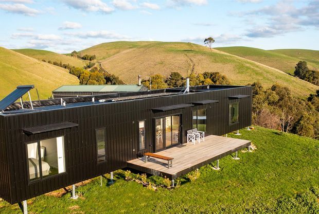 Aerial exterior view of a contemporary single-storey black-clad house set on a grassy elevated rural site. The elongated building has a low-pitched roof, extensive glazing, and a raised timber deck with outdoor seating. Rolling green hills and scattered trees form the surrounding landscape.