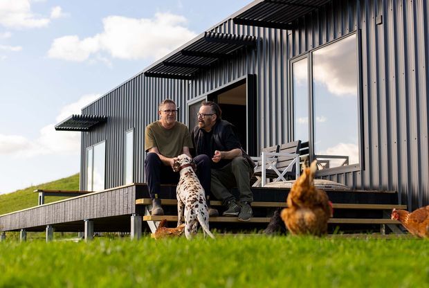 A contemporary single-storey building clad in dark vertically ribbed metal, set above a grassy site with a raised timber deck and steps. Two people sit outside with a dog, while chickens occupy the foreground. Large windows, an open glazed door, and projecting horizontal sunshades define the facade.