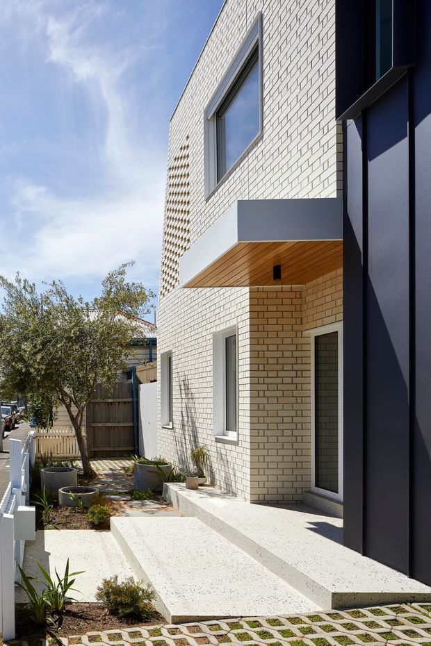 A contemporary multi-storey residential facade viewed from a narrow landscaped side path. The building combines pale cream brickwork with a dark charcoal vertical entrance volume, projecting box canopy, warm timber soffit, and slim grey-framed windows and door. Concrete steps and a ramp-like path lead to the entry, with raised planters, potted vegetation, a timber boundary fence, and permeable grass-filled pavers along the foreground.