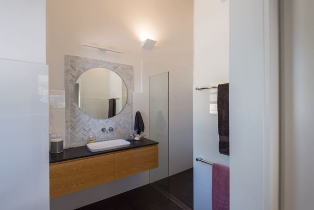 Bright contemporary bathroom with a wall-mounted timber vanity, dark stone benchtop, white vessel basin, large round mirror, and a patterned pale herringbone tile feature wall. A frameless glass shower enclosure and towel rails with dark and pink towels are visible. The room has a clean, refined, softly warm aesthetic.