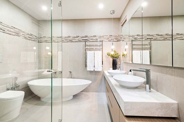 Bright contemporary bathroom with a freestanding white oval bathtub, glass-enclosed shower, wall-mounted toilet, double vessel basins, and a long pale stone vanity. Large mirrored cabinetry and a horizontal mosaic tile accent band create a clean, polished finish.