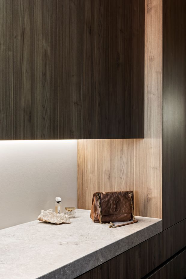 Close-up view of a refined built-in vanity or storage joinery installation. Dark timber-grain cabinetry contrasts with lighter timber vertical panels, a pale stone benchtop, and softly illuminated white textured wall detailing. A handbag and small decorative objects sit on the counter.