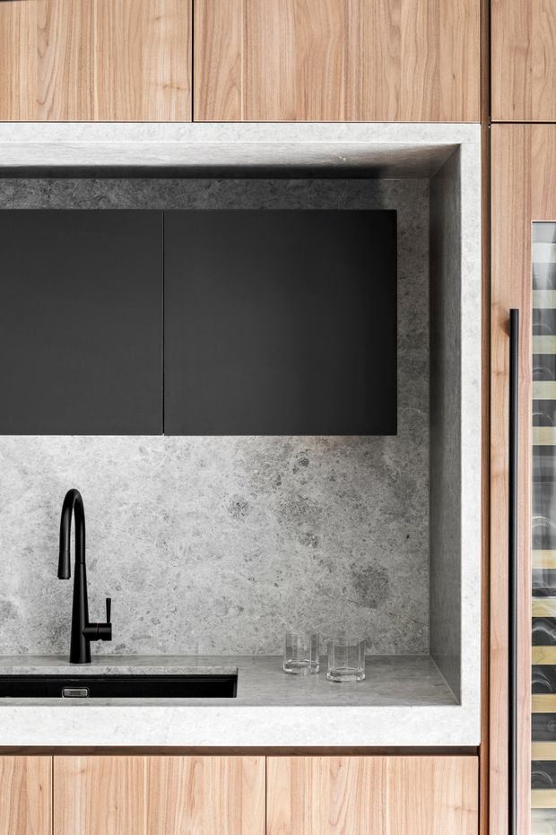 Close-up view of a refined built-in kitchen or bar joinery installation. Natural timber cabinetry frames a dark matte overhead cabinet and a grey textured stone or concrete-look recessed backsplash and benchtop. A black undermount sink and sculptural black tap provide strong contrast, while a narrow glass-front wine storage section is visible at the right edge.