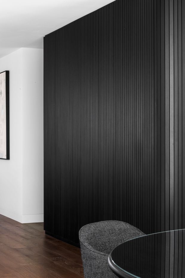 A refined interior corner featuring a dramatic full-height black vertical fluted wall or screen, contrasting white plaster walls and warm dark timber flooring. A curved grey upholstered chair and the edge of a round glass-topped table occupy the foreground, with a partially visible framed artwork on the left wall.