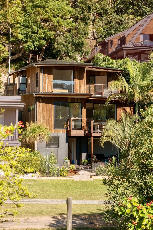 A multi-level timber-clad residential house set within lush, mature landscaping. The facade features extensive glazing, projecting balconies with horizontal metal balustrades, dark roof planes, and a mix of warm timber and dark painted wall surfaces. A lawn and garden path lead toward the lower-level entrance, while dense tropical and subtropical planting surrounds the building. The image is a bright exterior architectural photograph.