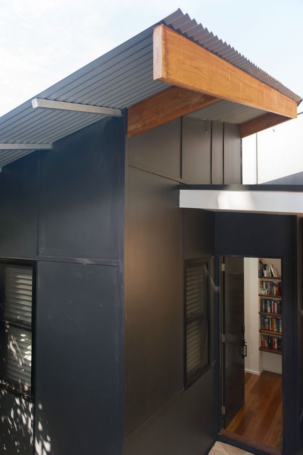 Close-up exterior architectural view of a compact modern structure with dark metal wall cladding, corrugated metal roofing, and prominent exposed warm timber roof beams and fascia. A dark-framed open doorway reveals a timber-floored interior with a tall built-in bookshelf. The composition highlights intersecting roof planes, projecting eaves, and crisp contemporary detailing.