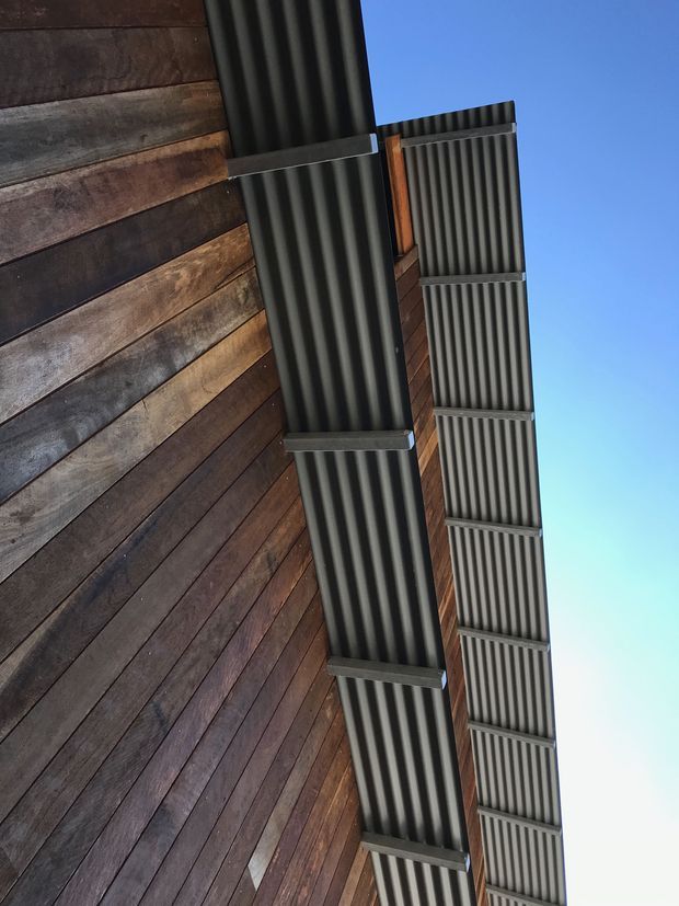 Upward-looking close-up of a building exterior showing dark stained horizontal timber wall cladding and projecting corrugated metal roof or canopy elements with dark metal framing. The image is dominated by the facade and roof undersides against a clear blue sky.