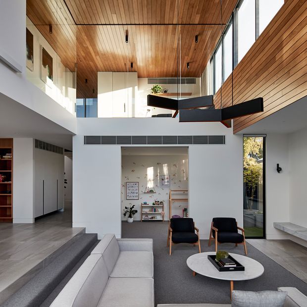 Double-height contemporary living area with a warm timber-lined ceiling and upper-level void. The room features a large light-grey sectional sofa, dark upholstered lounge chairs, a round coffee table, polished grey flooring, white walls, extensive glazing, and a dramatic black angular suspended pendant. Built-in cabinetry and a children's nook are visible at the rear, with a slim stair or landing connection at the upper level.