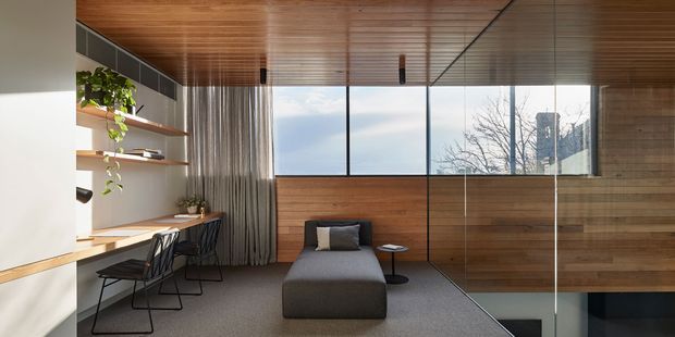 A compact contemporary living and workspace area with warm timber wall and ceiling lining, floor-to-ceiling glazing, built-in open shelving and desk, two black wire-frame chairs, and a dark upholstered lounge chair with ottoman. A glazed stair opening is visible at right, with daylight and trees seen beyond the windows.