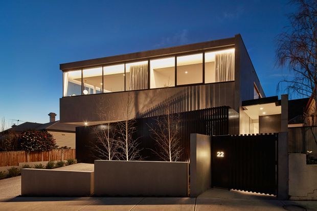 Twilight exterior view of a contemporary multi-level residence with a strong rectilinear form, dark metal screening and cladding, expansive upper-level glazing, warm recessed interior lighting, and a gated entry marked with the number 22. Low rendered planter walls and sparse deciduous landscaping frame the street frontage.