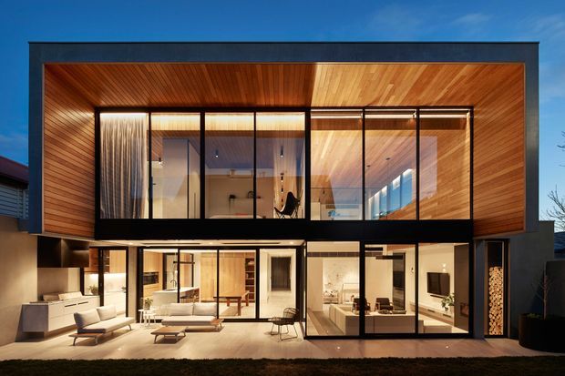 Contemporary two-storey residence viewed from the rear, featuring extensive floor-to-ceiling glazing, a projecting upper volume, warm timber soffits and wall cladding, dark-framed windows, and an illuminated open-plan ground floor with living and kitchen areas visible inside. The design creates a strong indoor–outdoor connection and a sheltered terrace-like outdoor living zone.