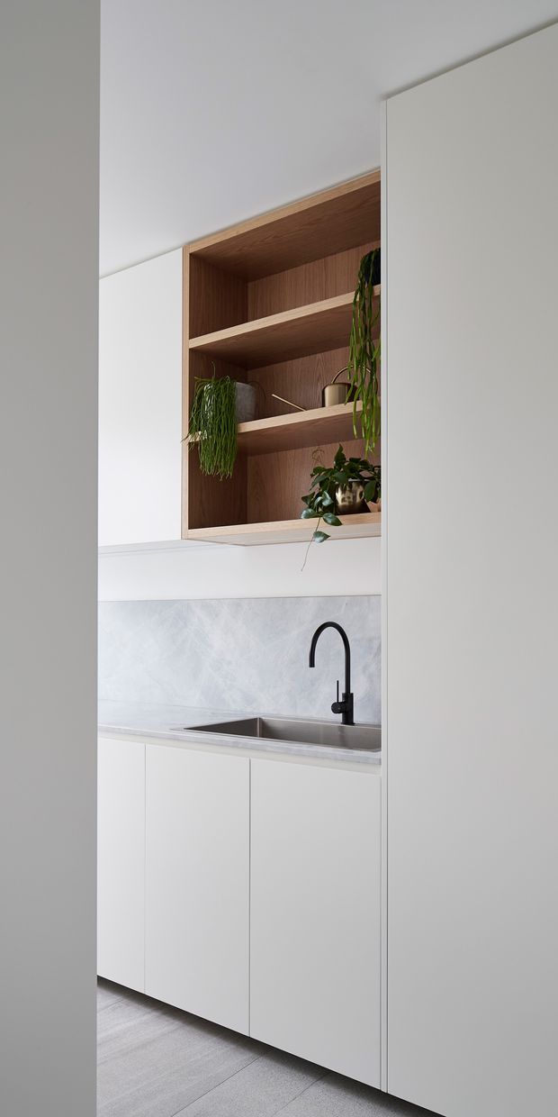 A compact, minimalist kitchen or kitchenette viewed through a doorway, featuring white flat-panel cabinetry, a stainless-steel undermount sink with a matte black gooseneck tap, a pale grey stone-like benchtop and splashback, and an open recessed timber shelving unit displaying potted plants and small objects. The room has a clean, bright, restrained palette with light grey flooring and warm timber joinery.