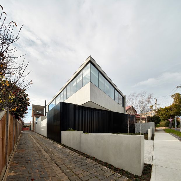 Street-level exterior view of a contemporary two-storey residential building on a suburban corner site. The sharply angled upper volume has extensive floor-to-ceiling glazing, pale rendered or concrete-like wall surfaces, and a dark vertical ribbed facade at ground level. A low light-coloured masonry boundary wall, paved laneway, driveway, neighbouring homes, and mature street vegetation are visible.