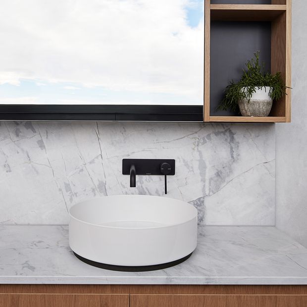 Contemporary bathroom vanity with a white round vessel basin on a grey-white marble-look benchtop and splashback. A matte black wall-mounted tap set, black-framed mirror, and integrated timber display niche with a potted plant provide contrast. Timber cabinetry is visible below the vanity.