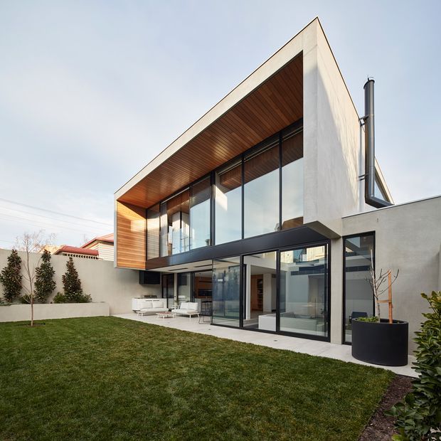 Exterior view of a contemporary two-storey home with a sharply angled roofline, projecting upper-level volume, extensive floor-to-ceiling glazing, dark metal window frames, white rendered walls, and a warm timber-lined soffit and facade. The house opens onto a manicured lawn and enclosed courtyard-like garden, with outdoor seating visible beneath the upper level.