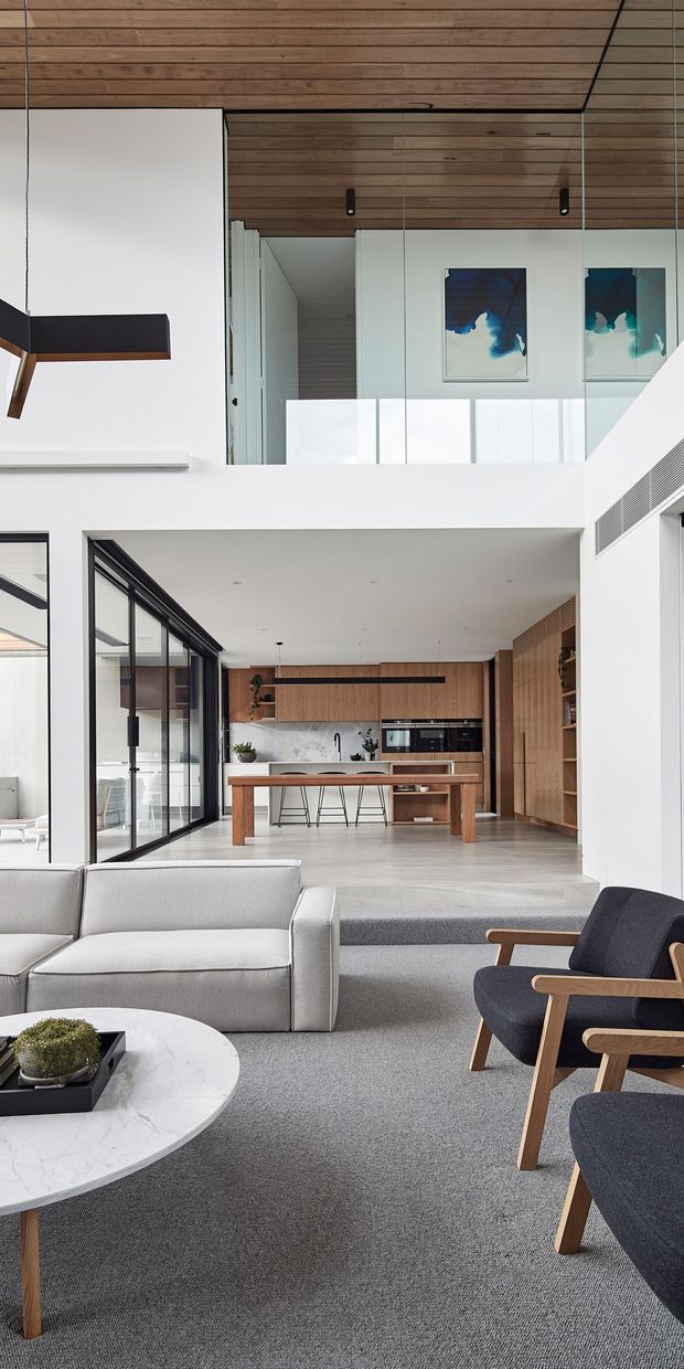 A bright, open-plan contemporary living room looking through to a timber-lined kitchen. The foreground contains a light grey sofa, navy upholstered timber-framed armchairs, a round marble coffee table, and grey carpet. A raised kitchen zone features timber cabinetry, a marble-look splashback, central island, integrated appliances, and black-framed glazed doors. A double-height void with a white balustrade, timber ceiling, internal glazing, and framed artwork adds architectural interest.