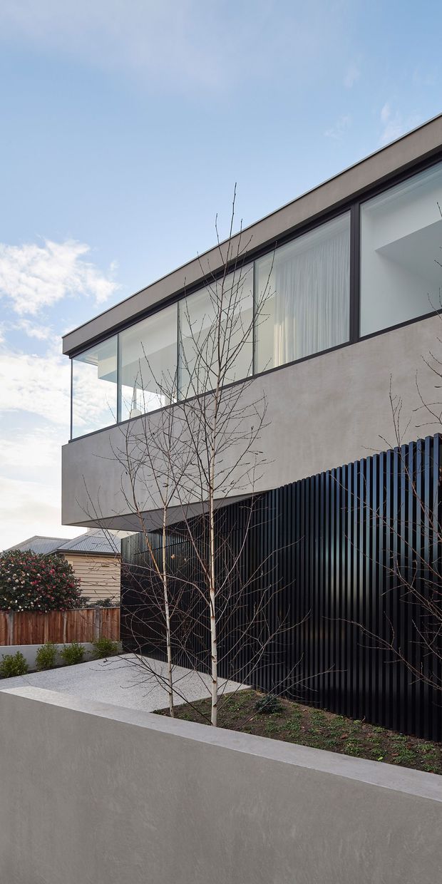 Exterior photograph of a contemporary two-storey residence in a suburban setting. The building has a minimalist rectilinear form, smooth light-grey rendered walls, extensive dark-framed glazing, a cantilevered upper level, and a black vertical slatted facade or privacy screen. A paved front garden with young deciduous trees and a low rendered boundary wall occupies the foreground.