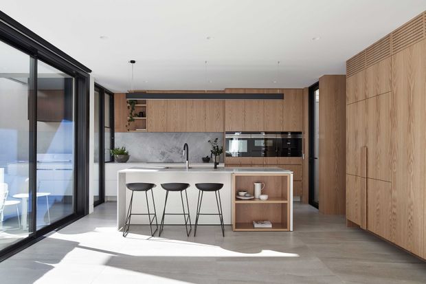 Bright contemporary kitchen with a large central island and three black bar stools. The space features extensive warm timber cabinetry, integrated appliances, open shelving, a white benchtop, dark-framed floor-to-ceiling glazing, and grey-toned flooring. Strong natural light creates a clean, refined, open atmosphere.