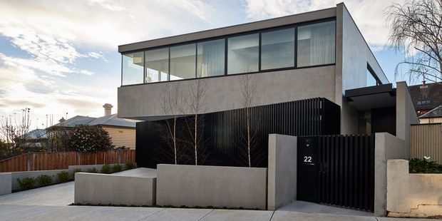 Street-facing exterior of a contemporary multi-level residence with a flat roof, strong horizontal geometry, extensive upper-level glazing, a rendered concrete-toned facade, and a black vertical slatted privacy screen and entry gate. The property includes a paved forecourt, low rendered planter walls, timber fencing, and neighbouring suburban homes.