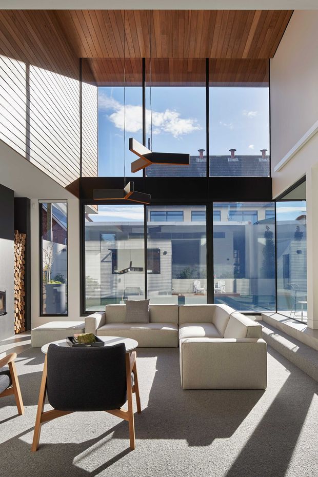 A bright, double-height contemporary living room with a large L-shaped upholstered sectional, armchairs, and a low coffee table. Floor-to-ceiling glazing opens views toward an outdoor pool courtyard and neighbouring buildings. The room features a warm timber-lined ceiling, extensive black-framed glazing, pale walls, carpeted flooring, and sculptural modern pendant lights. Strong sunlight creates long geometric shadows across the room.