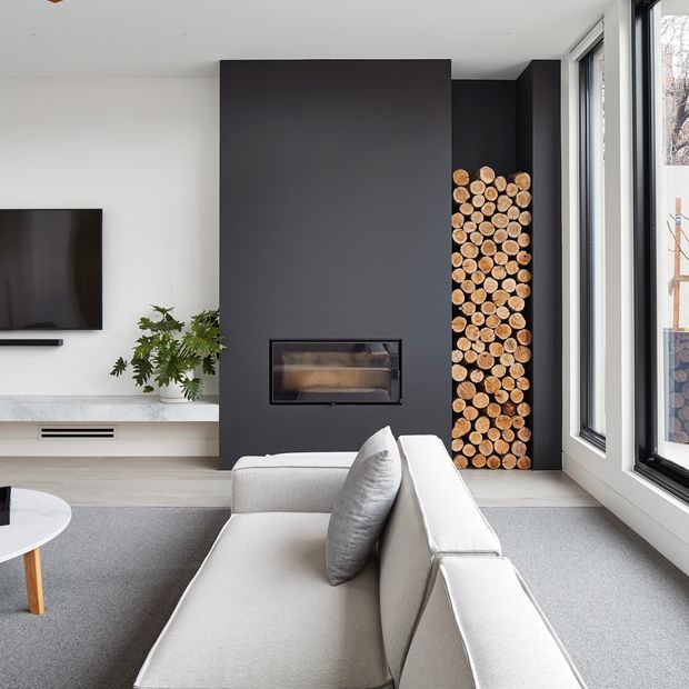 Bright contemporary living room featuring a large light upholstered sectional sofa, a dark charcoal fireplace feature wall with a built-in fireplace, a neatly stacked firewood niche, wall-mounted television, pale stone or concrete-look media console, broad grey carpet, and full-height black-framed glazing.