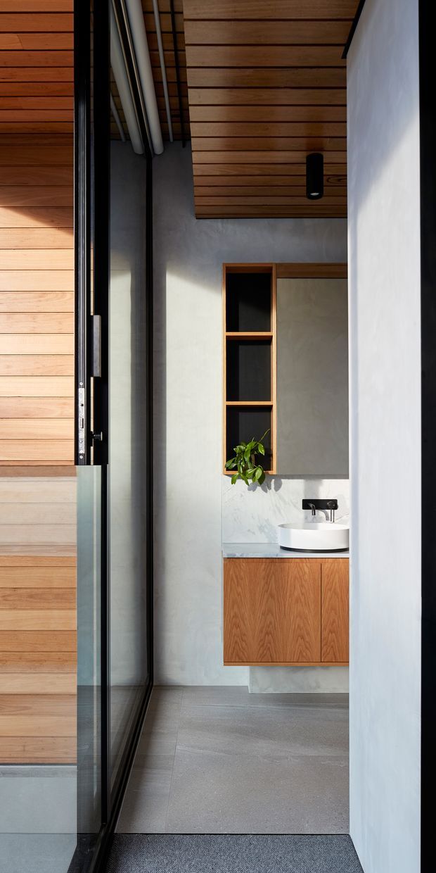 A compact contemporary bathroom viewed through a dark-framed glazed entry. The space features a floating warm timber vanity with a white round vessel basin, black wall-mounted tapware, a large frameless mirror, open timber shelving with a plant, smooth pale grey walls, grey tiled flooring, and a horizontal timber-lined ceiling. Strong vertical glazing and clean built-in detailing create a refined indoor-outdoor threshold.