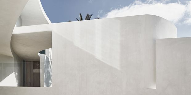Close-up exterior architectural view of a sculptural white rendered building with smooth curved walls, sweeping roof or canopy forms, deep reveals, and dark glazed openings. Palm fronds and blue sky are visible above, suggesting a sunny contemporary setting. The composition emphasises minimal geometric forms and strong light-and-shadow contrasts.