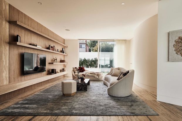 A refined contemporary living room with a long timber feature wall incorporating built-in open shelving and a wall-mounted television. Neutral curved upholstered seating surrounds a dark coffee table, with a large textured rug over timber flooring. Full-height rear glazing provides natural light and views to greenery, while recessed ceiling lights and pale walls create a calm, spacious atmosphere.