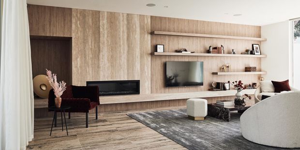 Contemporary living room featuring a full-height timber-toned feature wall with integrated fireplace and television, extensive open shelving, timber-look flooring, neutral upholstered seating, a dark area rug, and restrained decorative styling. Floor-to-ceiling curtains and recessed ceiling lights contribute to a refined, warm interior.