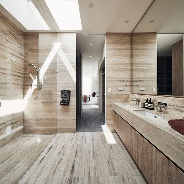 Bright contemporary bathroom with extensive light natural stone-look wall and floor finishes, a frameless glass shower, freestanding or partially visible bathtub, and a long timber-toned double vanity with an integrated basin and large mirror. A carpeted bedroom or dressing area is visible through the central opening, while skylights and recessed ceiling lights provide strong natural and artificial illumination.