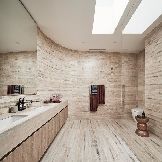 Contemporary bathroom with curved walls clad in pale veined stone-look tile, matching flooring, a long floating timber vanity with integrated white basin, large wall mirror, skylights, and a wall-mounted toilet. Dark towels and small accessories provide contrast against the warm neutral finishes.
