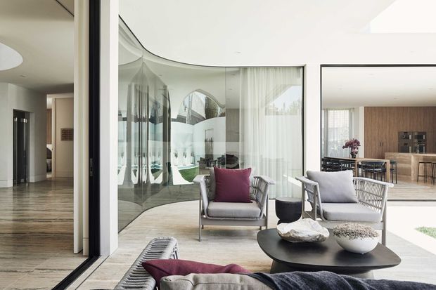 Bright contemporary open-plan interior with a sophisticated living room at the centre, featuring two light upholstered lounge chairs, a low black organic-shaped coffee table, and a neutral sofa. Curved full-height glazing and mirrored surfaces create a layered connection to the courtyard or exterior. A dining area and timber kitchen with an island are visible beyond, while a polished stone or concrete-toned floor extends through the space. The palette is predominantly white, light grey, beige, and timber, with a deep plum accent cushion.