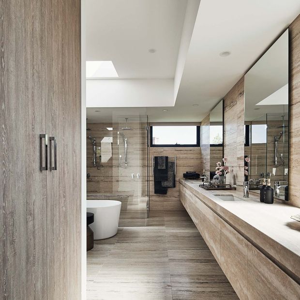 Contemporary bathroom with a long double vanity, wall-to-wall mirror, floating timber-look cabinetry, large-format timber-look tiled flooring and wall finishes, a glass-enclosed shower, freestanding white toilet, recessed lighting, and a skylight. The palette is warm neutral beige, taupe, and light wood tones with black hardware accents.
