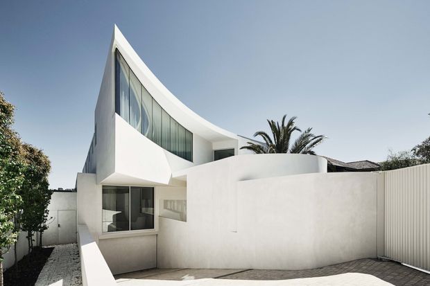 Exterior view of a sculptural contemporary white house with a sweeping curved roofline, large floor-to-ceiling glazed windows, smooth rendered walls, and a private courtyard or driveway enclosure. Palm and garden planting are visible around the property.