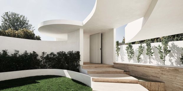 A sculptural modern residential entrance with sweeping curved white concrete forms, a tall grey entrance door, warm timber-clad steps, and integrated curved garden walls. The composition is bright and minimalist, with strong interplay between architectural curves, shadows, and landscaped planting.