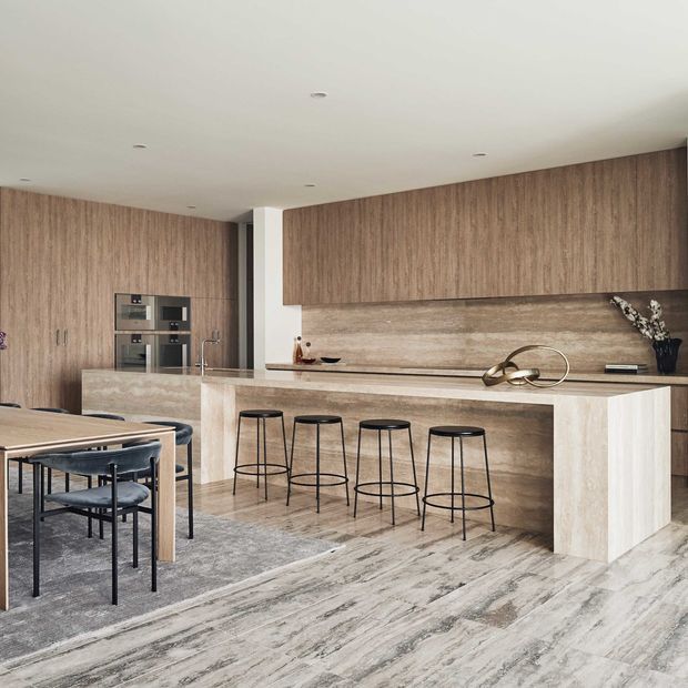 Contemporary open-plan kitchen and dining area with extensive warm wood cabinetry, a large stone island with four black bar stools, integrated appliances, and a dining table positioned on a grey rug. The space has a clean, refined look with a neutral palette and dramatic veined flooring.