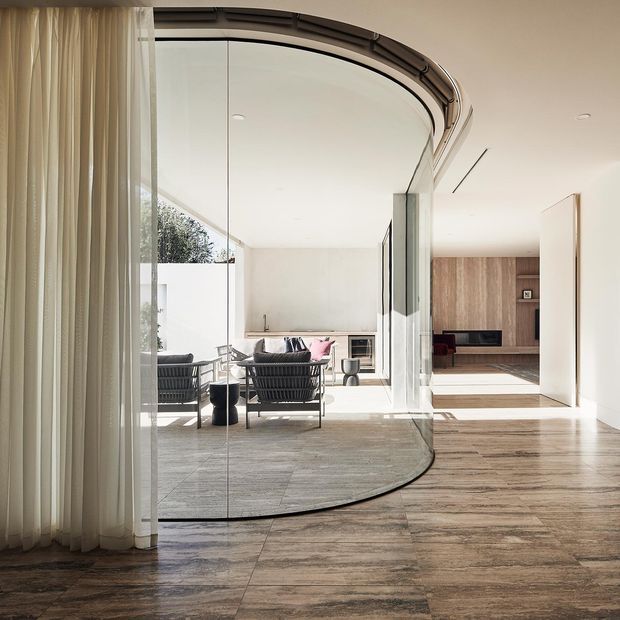 Bright contemporary living area featuring a dramatic curved glazed wall and matching curved ceiling track, sheer floor-to-ceiling curtains, and large wood-look tiled flooring. A sitting area with black lounge chairs and a sofa opens toward an exterior courtyard or garden. The room connects to a warm timber-lined media or living zone through a wide opening, with abundant natural light and clean white surfaces.