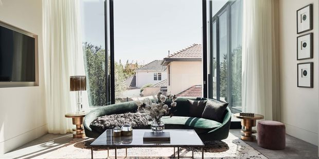 Bright, refined living room with a deep green curved sofa positioned beside full-height black-framed glazing and sheer curtains. The room includes a wall-mounted television, paired metallic side tables, a low rectangular coffee table, a patterned area rug, framed monochrome artwork, and a small upholstered ottoman. Sunlight and views toward neighbouring rooftops create an airy urban residential setting.